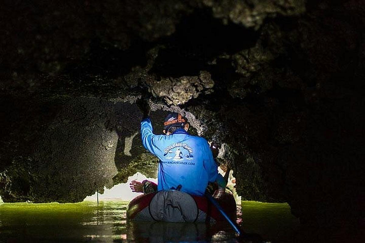 Person in a small boat navigating through a dark, narrow cave passage with overhead rock formations and water below.