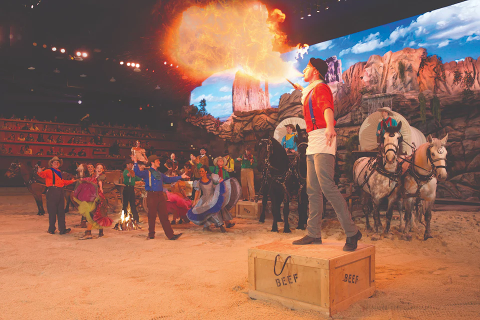 A performer breathes fire in a Western-themed show with costumed actors, horses, and a scenic desert backdrop.