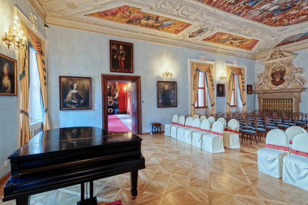Elegant room with ornate ceiling, wall paintings, chandelier, a grand piano, and a row of white chairs against the wall.
