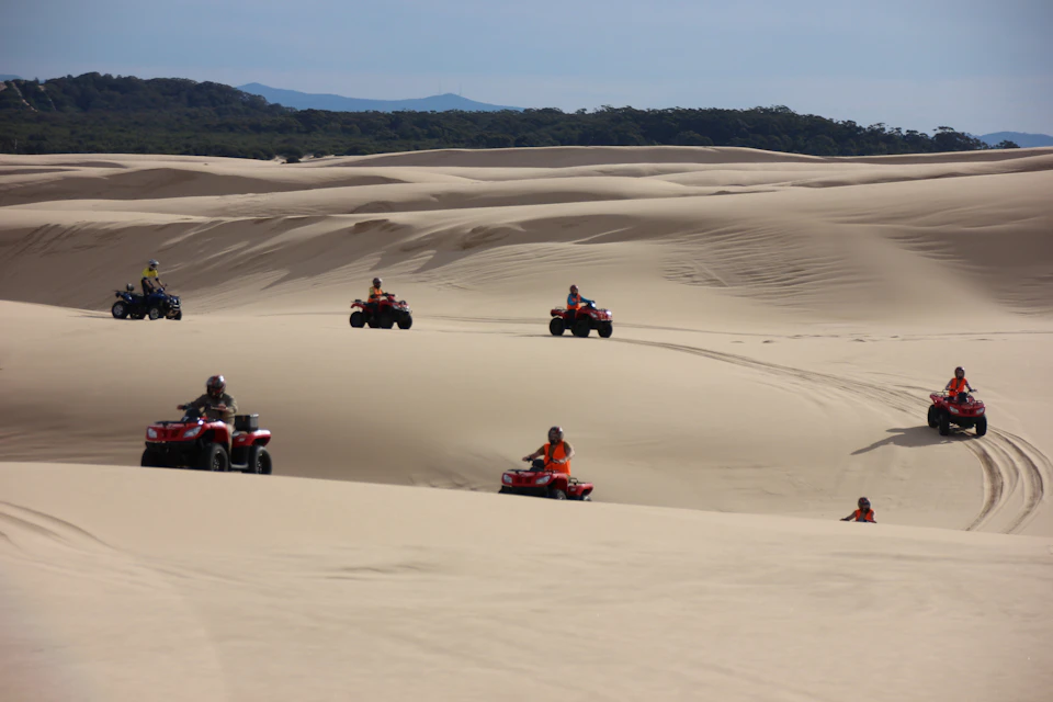 People riding red ATVs across vast sandy dunes under a clear sky. Forested hills are visible in the distance.