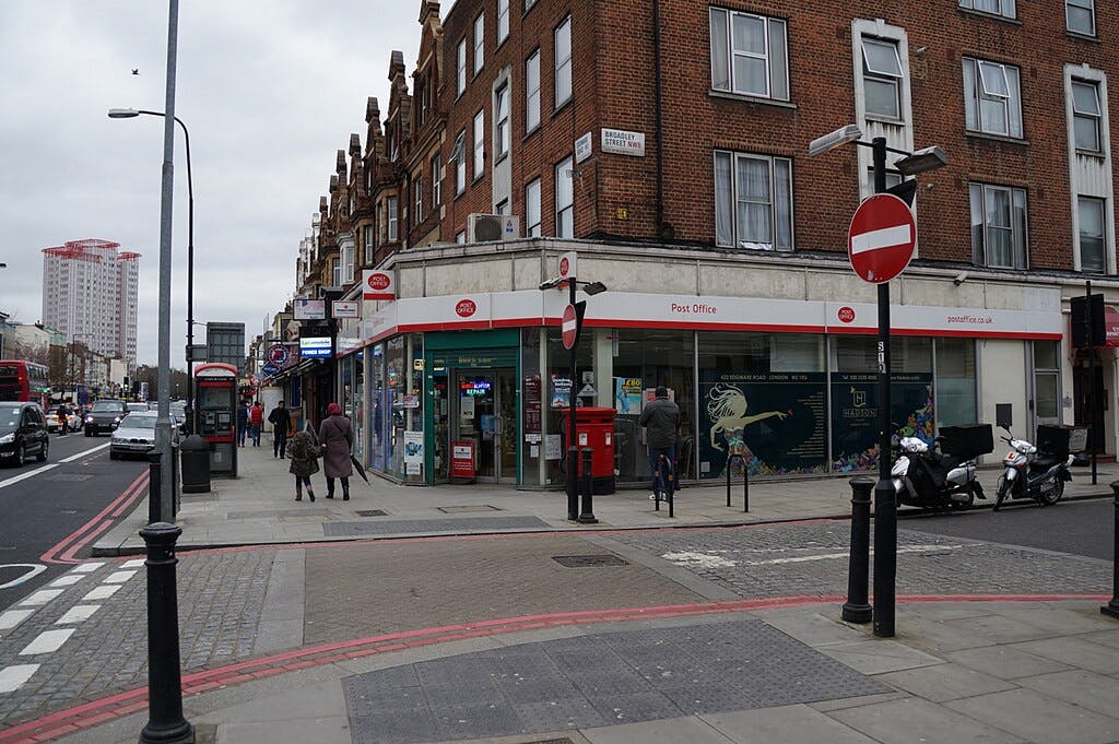 Urban street scene with a red-brick post office, pedestrians, parked scooters, a red phone booth, and nearby traffic.