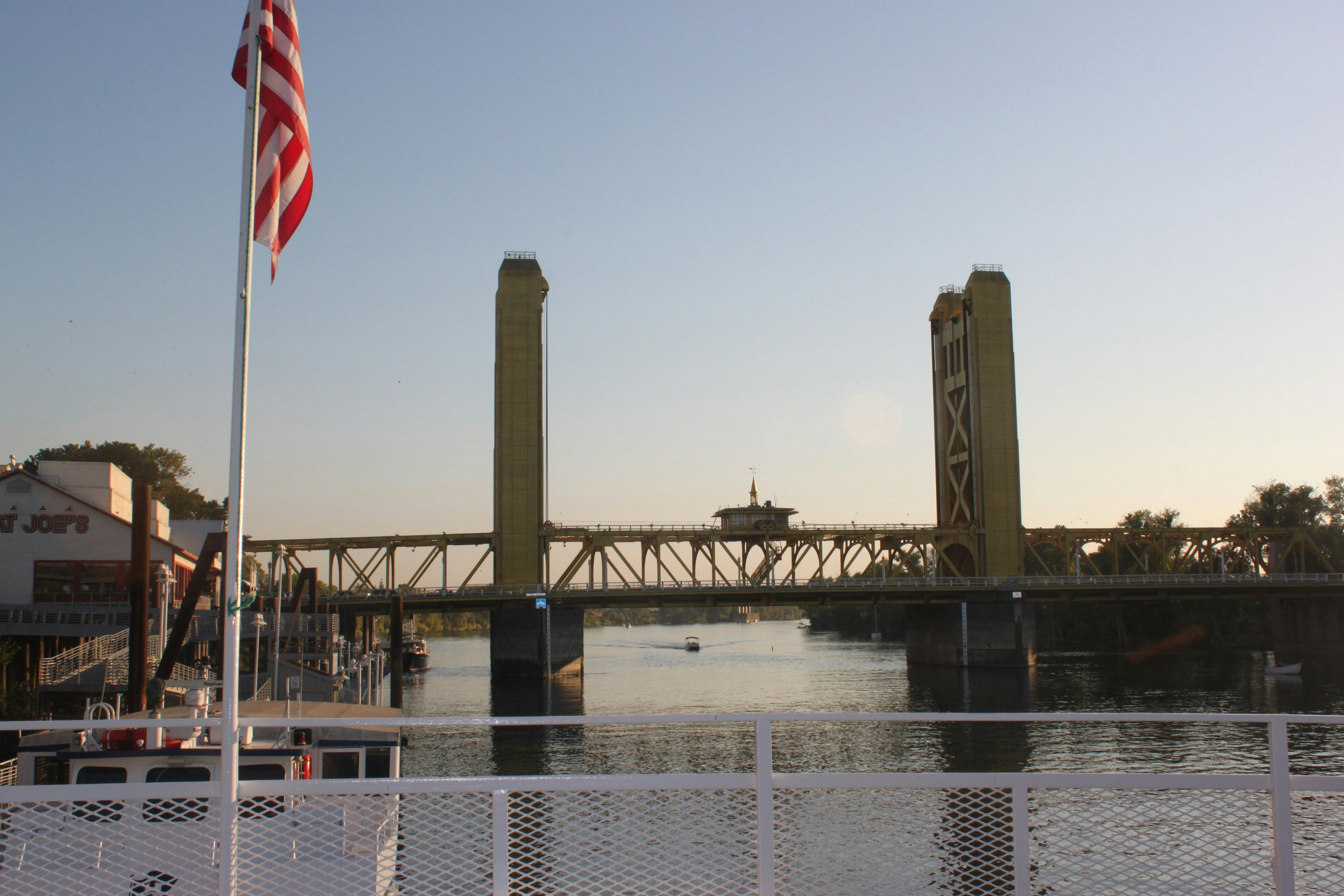 日落时分，一座高耸的吊桥横跨一条平静的河流。 前景中可以看到一面美国国旗。