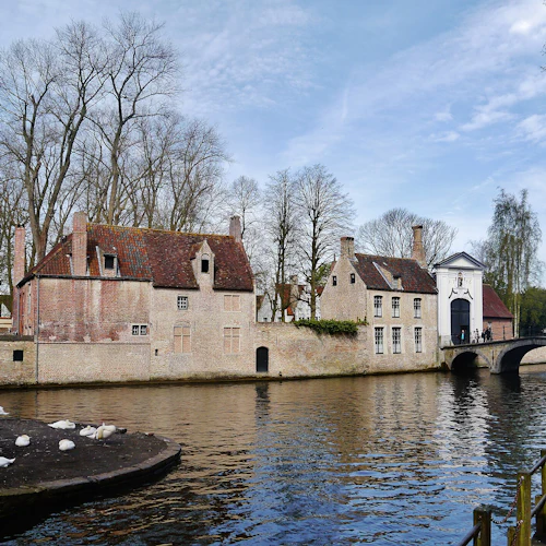Stenhus och en bro vid en kanal, med svanar på en ö i vattnet. Två personer som tar foton på ett trädäck.