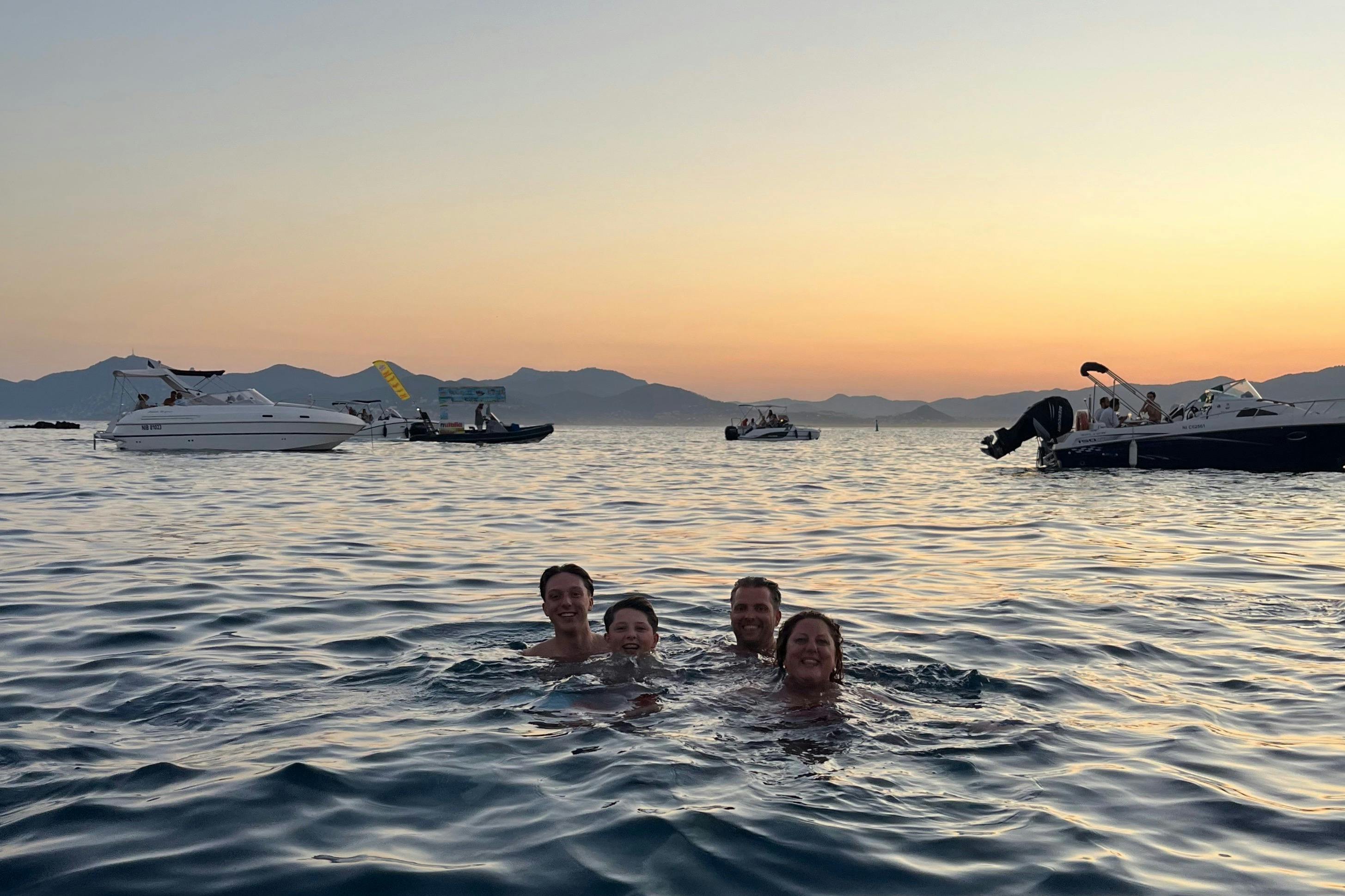 Four people swimming in the sea at sunset, with boats in the background and distant mountains visible.