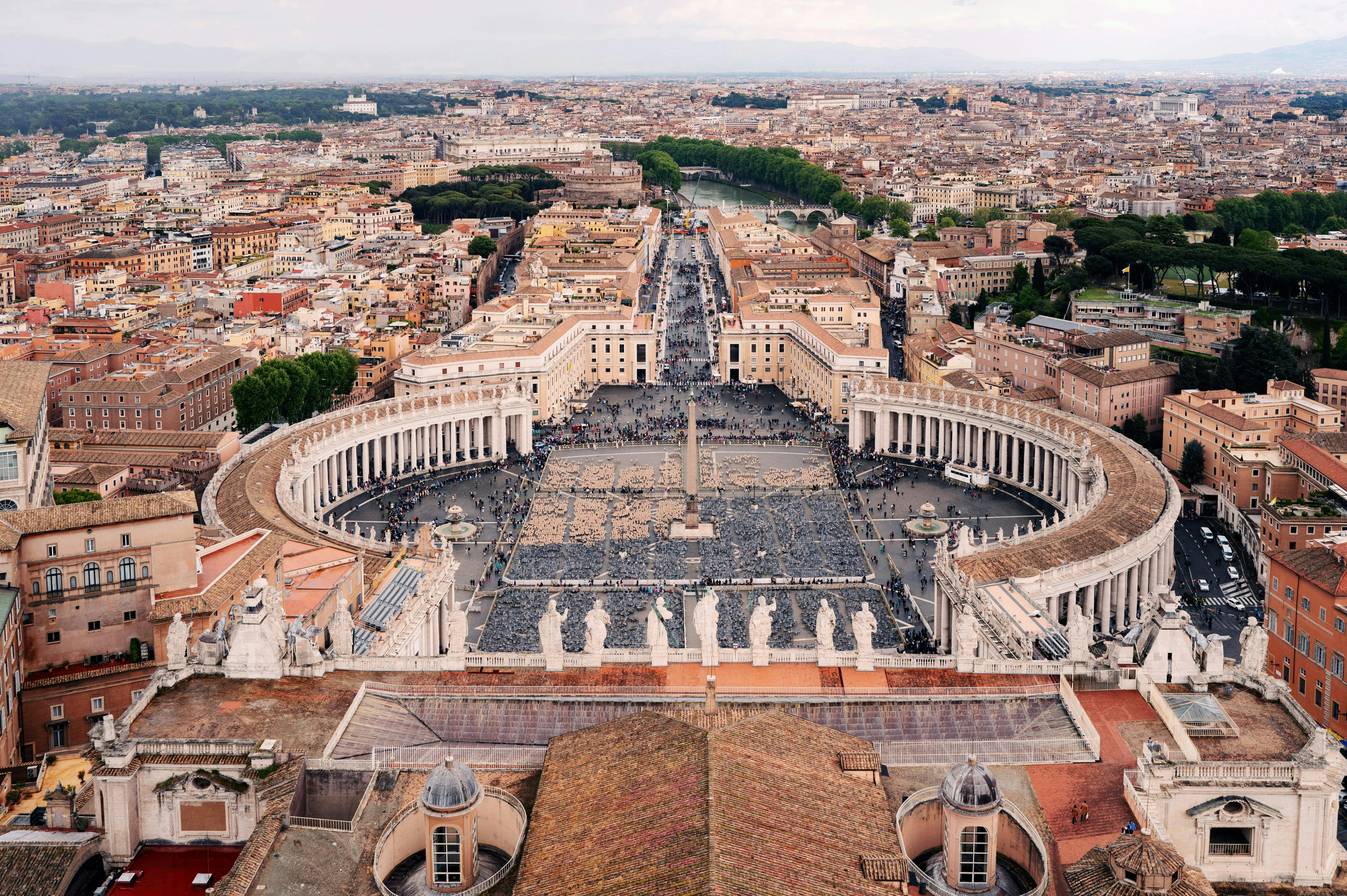 Vista aèria de la plaça de Sant Pere a la Ciutat del Vaticà, envoltada de columnates i estàtues, amb un fons de paisatge urbà llunyà.
