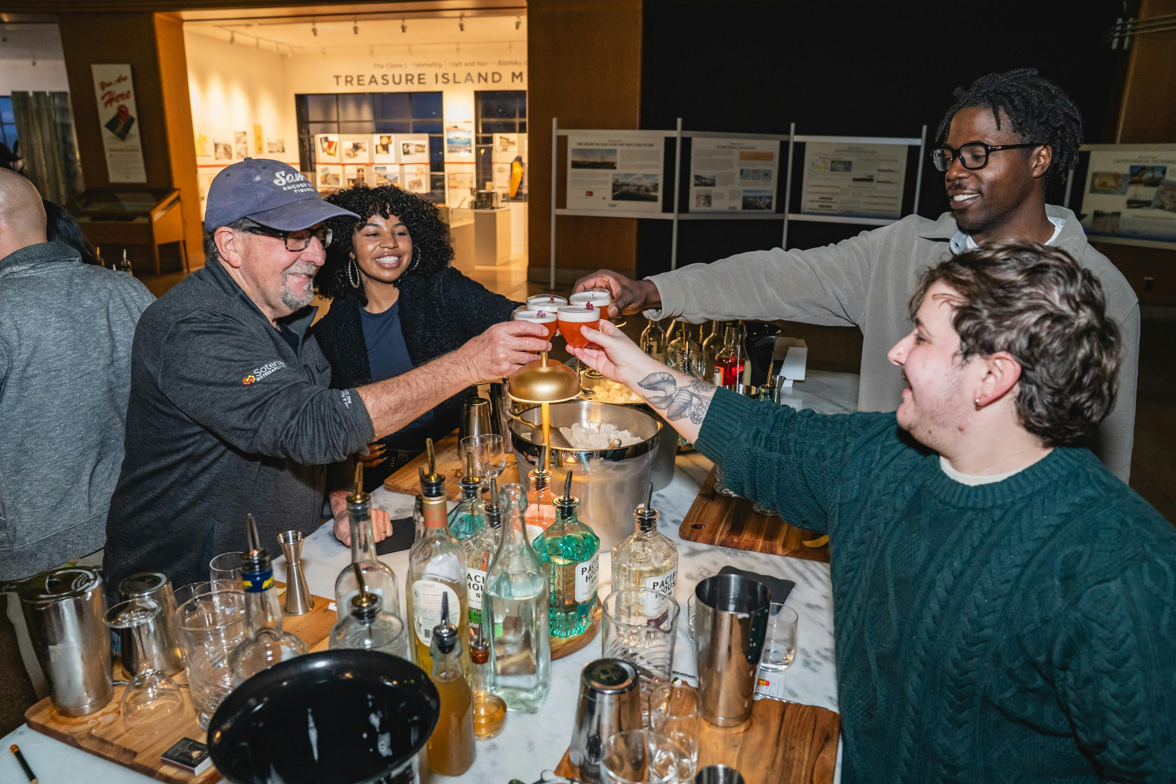 Four people standing around a bar counter cheer with glasses, surrounded by liquor bottles and bartending tools.