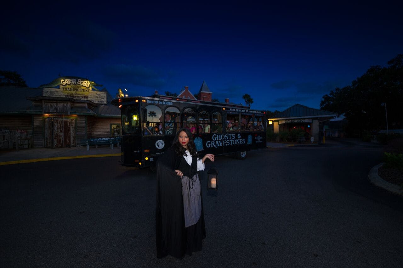A person in old-fashioned attire holds a lantern beside a black trolley with "Ghosts & Gravestones" written on it, at night.