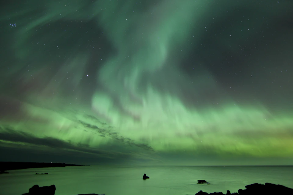 Green aurora borealis lights up the night sky over calm sea with scattered rocks; starry sky visible.