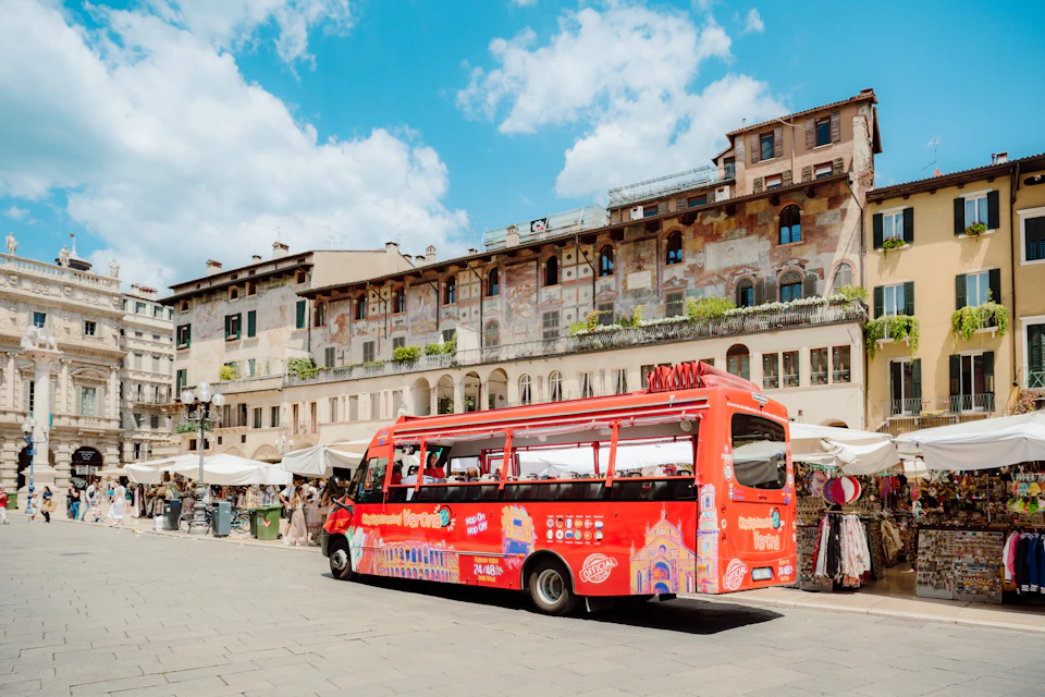 A red tour bus parked in a sunny plaza with historic buildings and market stalls in the background.