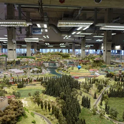 Miniatyr landskapsdiorama med träd, byggnader, vägar och en flod, sedd av flera personer i ett stort, väl upplyst rum.