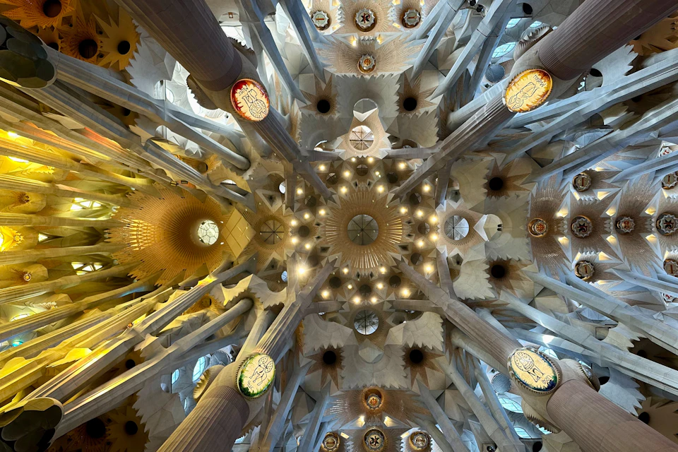 The intricate ceiling of a cathedral, showcasing symmetrical columns, ornate lighting, stained glass, and geometric patterns.