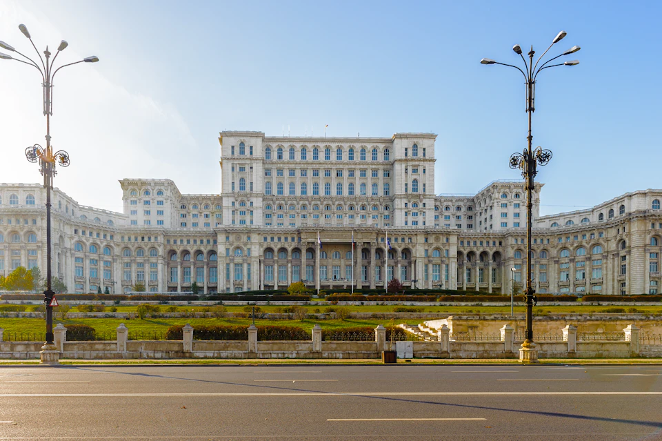A large, neoclassical building with numerous windows and columns, surrounded by landscaped gardens, seen from across a street.