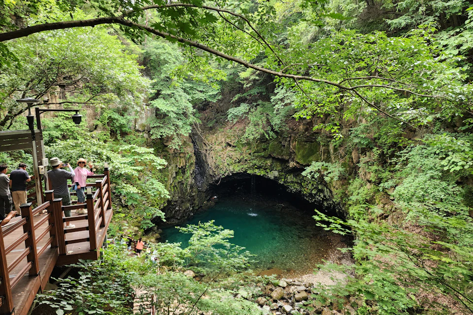 A lush green forest surrounds a small, clear turquoise pool at the base of a rocky cave. People are viewing from a wooden platform.