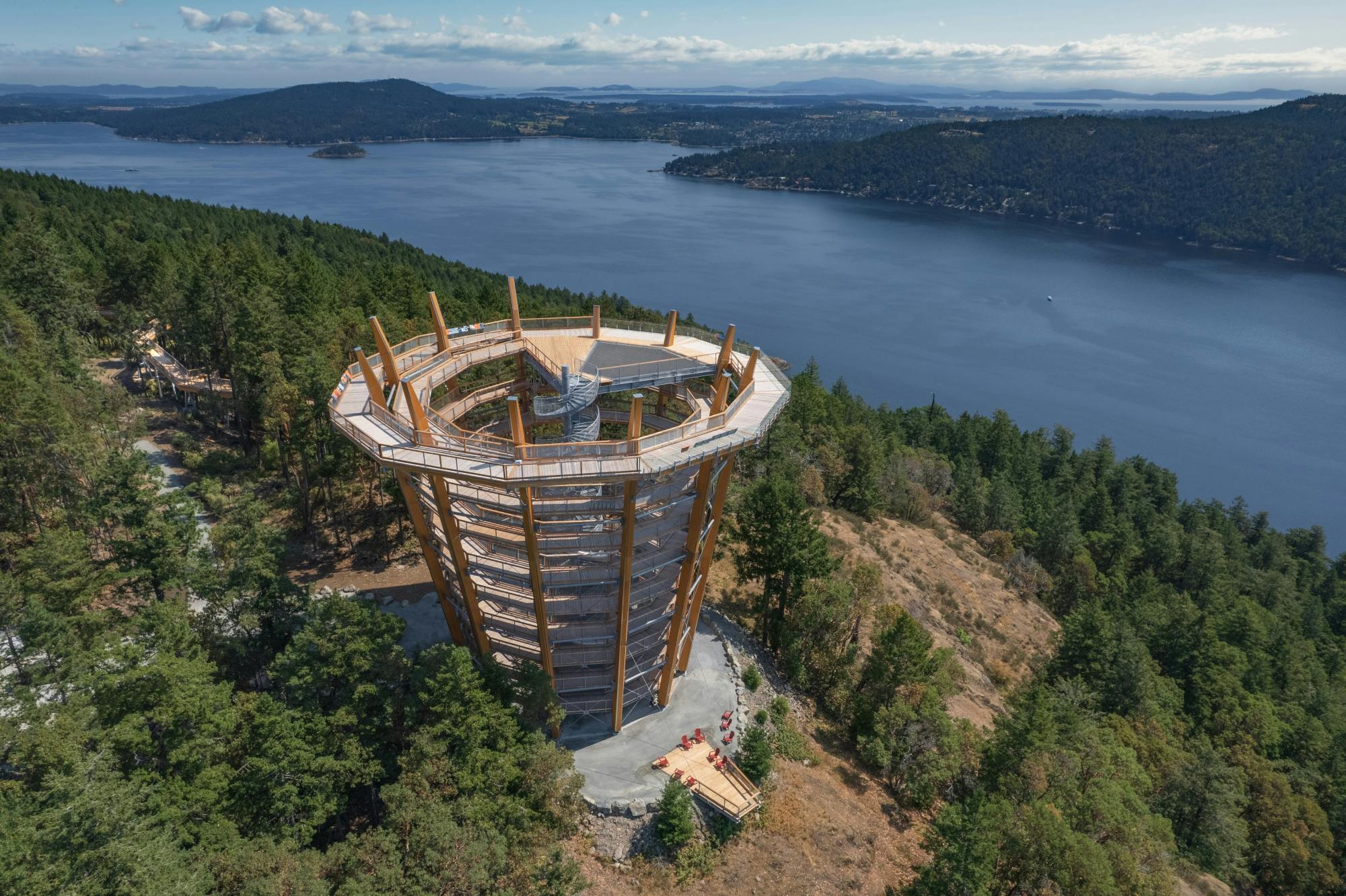 Observation tower with spiral walkway among dense trees, overlooking a serene body of water with hills in the distance.