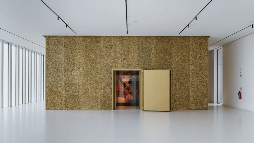 A minimalist room with a golden wall featuring an open door, revealing a colorful interior with a blurry figure and objects inside.