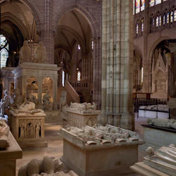 En historisk krypta med utsmyckade stensarkofager och skulpturer i en katedral, omgiven av glasmålningar och valv.