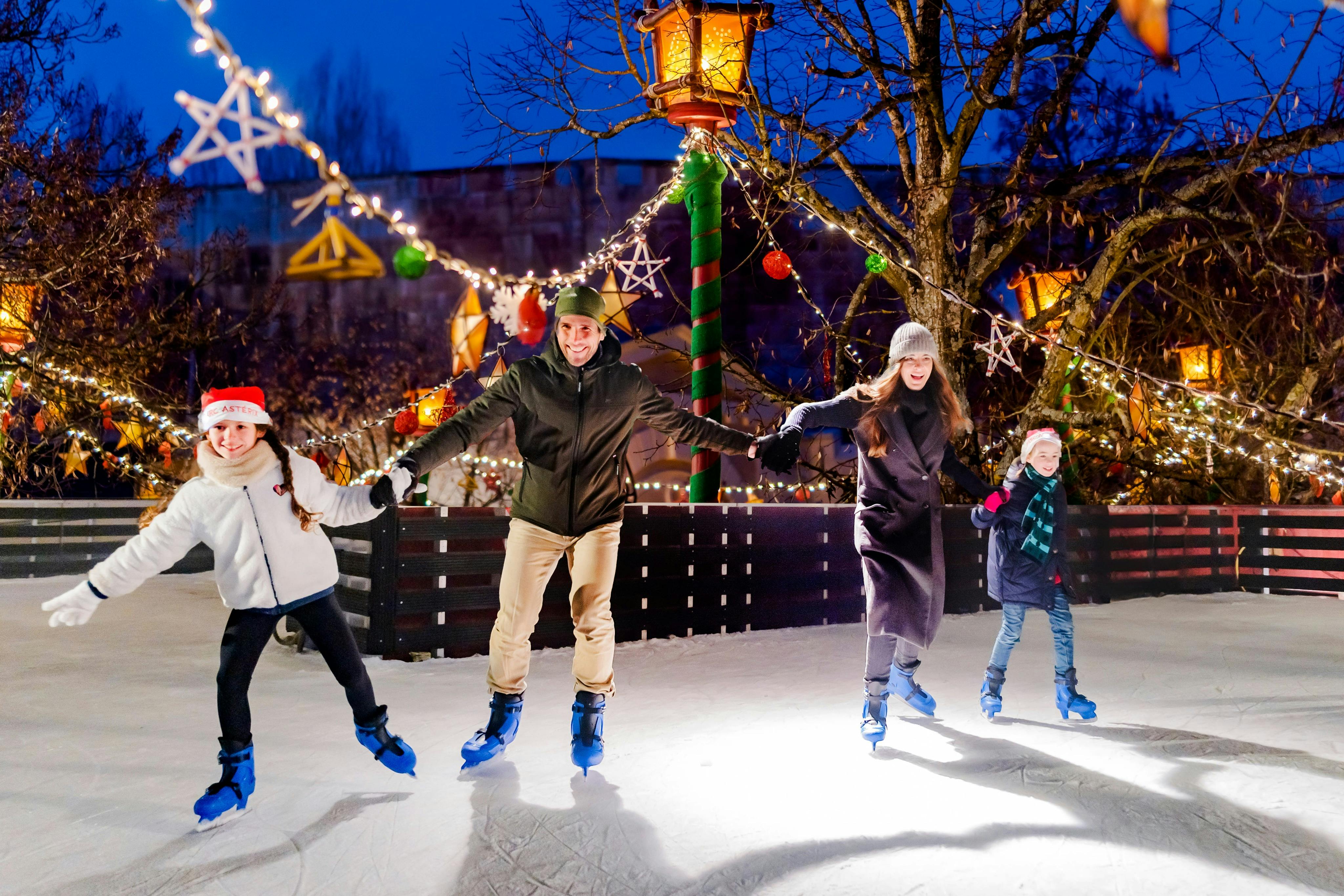 Three people ice skating outdoors at night under festive lights and decorations, with a cheerful atmosphere.