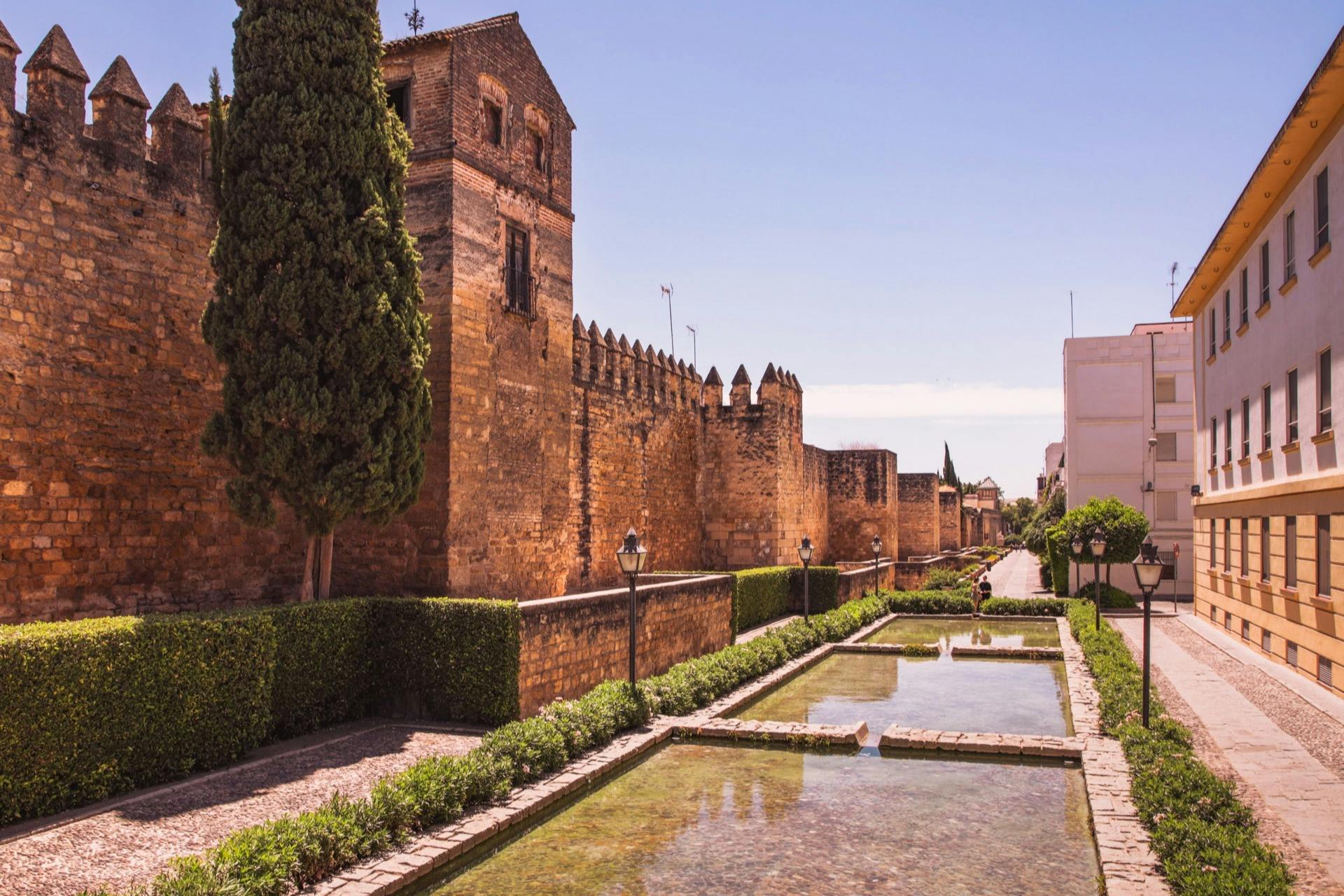 Historic stone fortress with crenellated walls, flanked by tall trees and a clear reflecting pool, under a clear blue sky.