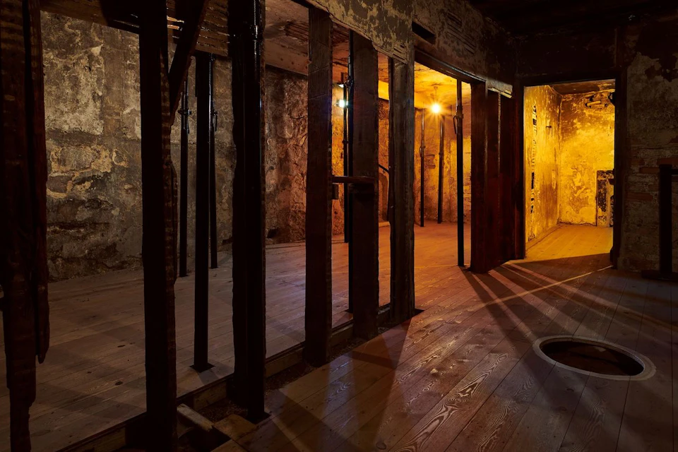 Dimly lit, rustic wooden room with exposed beams, stone walls, and warm lighting. No people are present.