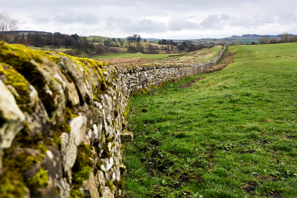 A long, moss-covered stone wall stretches across a green, rolling landscape under a cloudy sky.