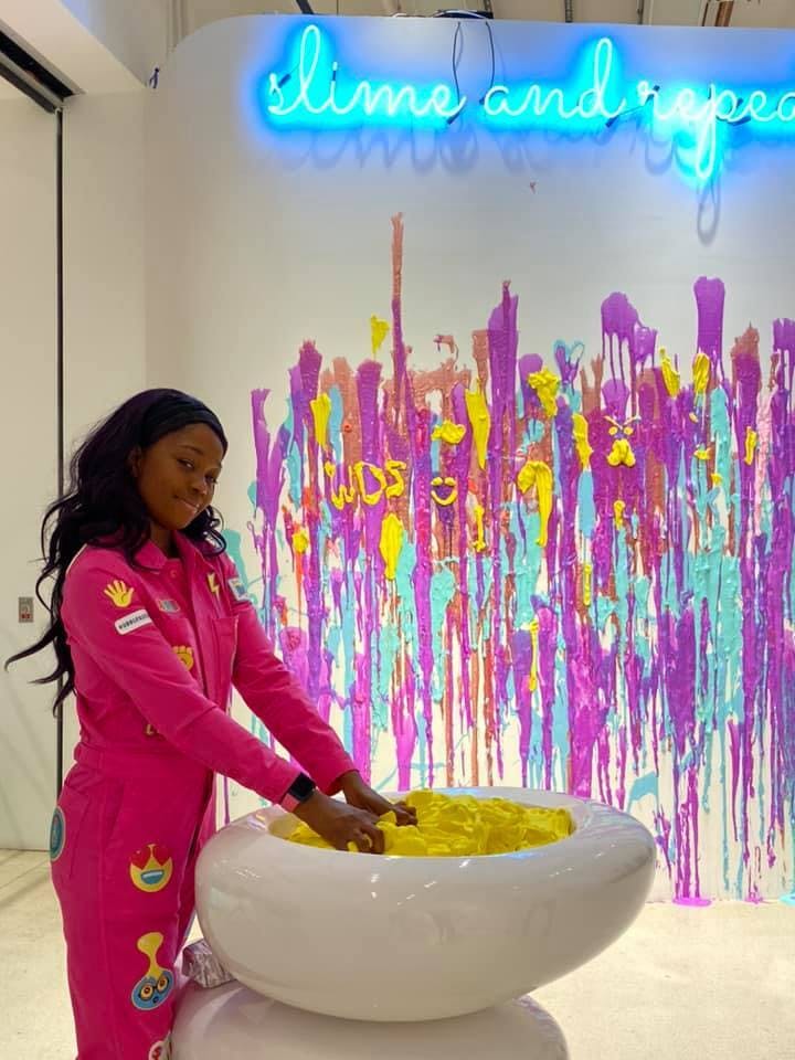 A woman in a pink jumpsuit interacts with a yellow substance in a white vessel, against a colorful, dripped paint wall with neon text above.
