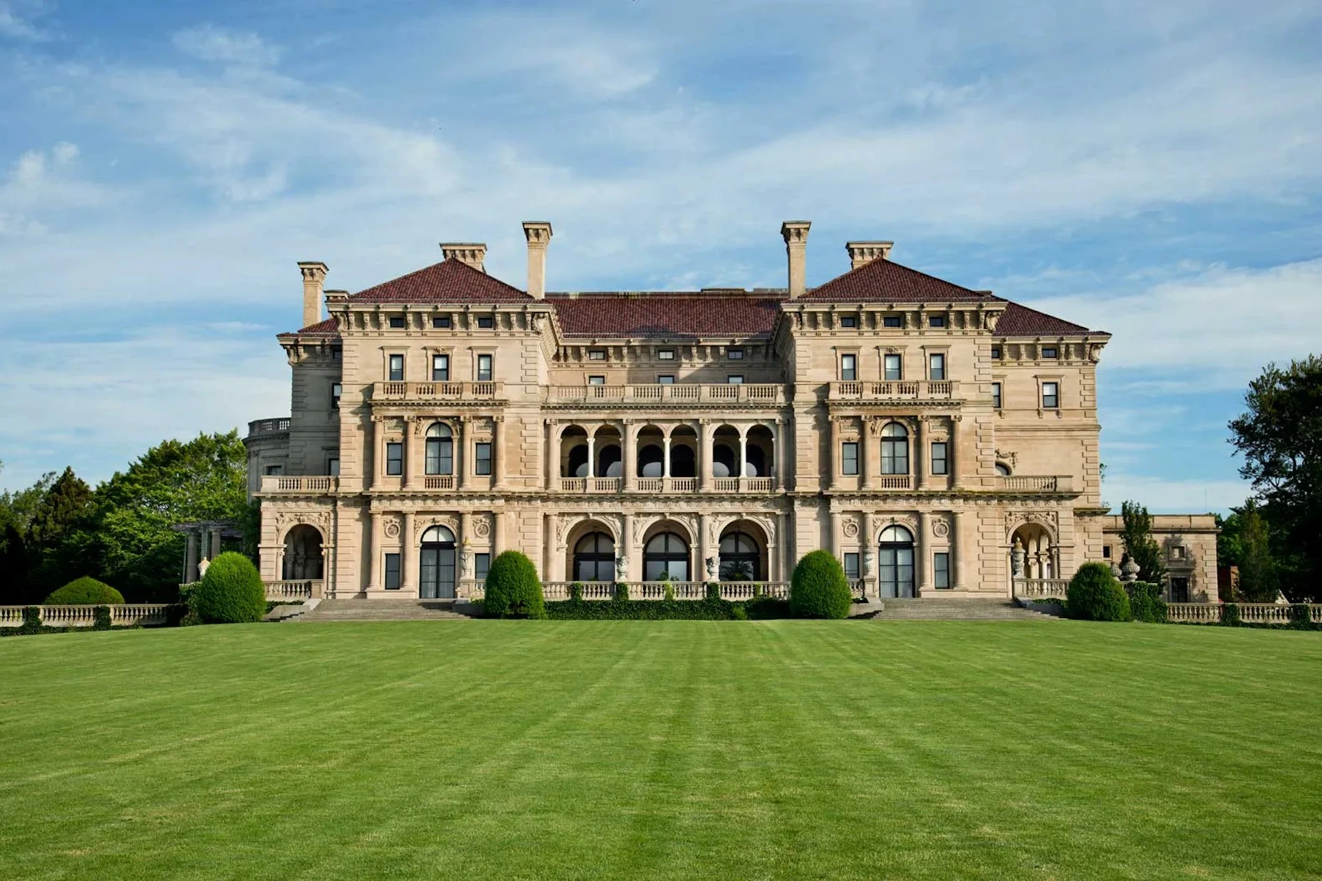A large, ornate mansion with symmetrical architecture, arched windows, and a well-manicured lawn under a blue sky.