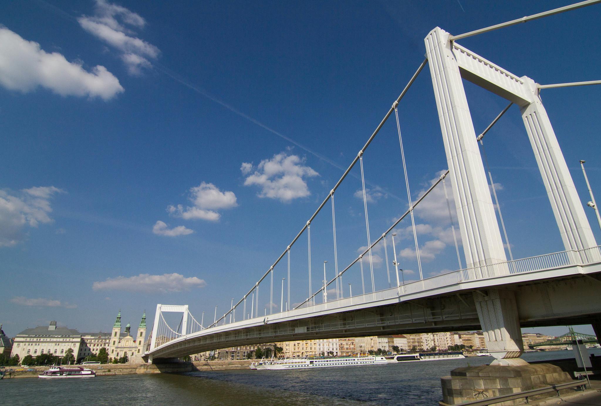 En vit hängbro sträcker sig över en flod under en klarblå himmel med spridda moln. Båtar och stadsbyggnader syns.