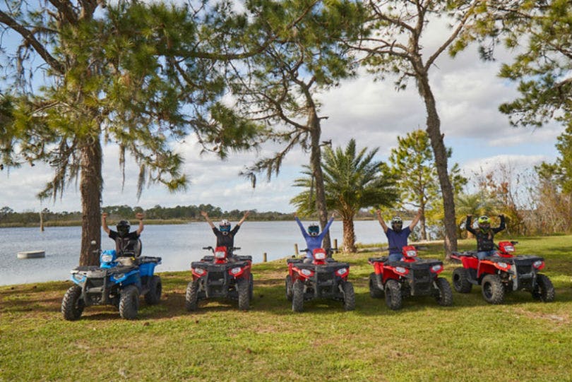 ATV를 탄 사람들은 팔을 들어 호숫가 잔디밭에 주차하고, 나무와 흐린 하늘을 배경으로 달립니다.