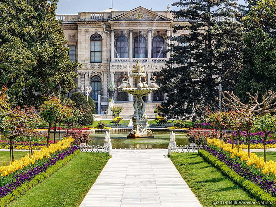 A historic building with arched windows, a landscaped garden, a central pathway, and an ornate fountain with sculptures.