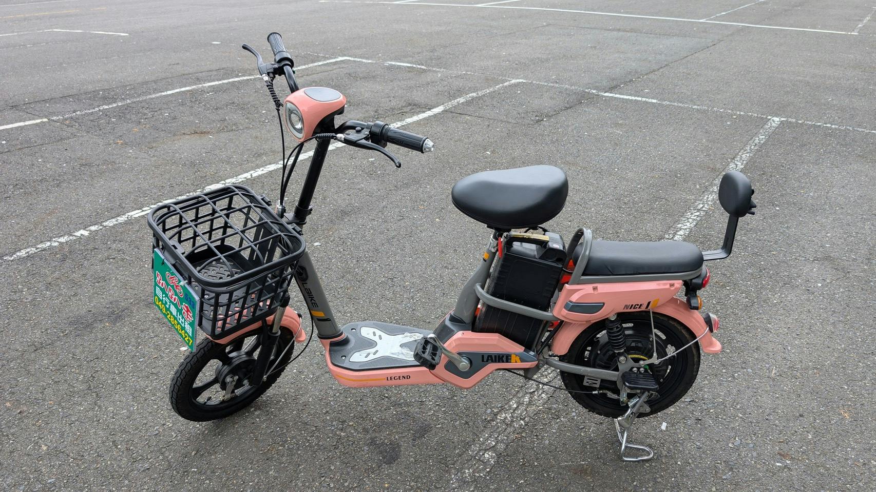 A pink electric scooter with a black seat and basket, parked in an empty lot with white parking lines visible.