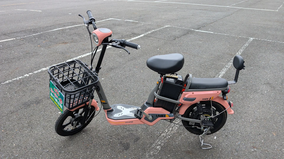A pink electric scooter with a black seat and basket, parked in an empty lot with white parking lines visible.