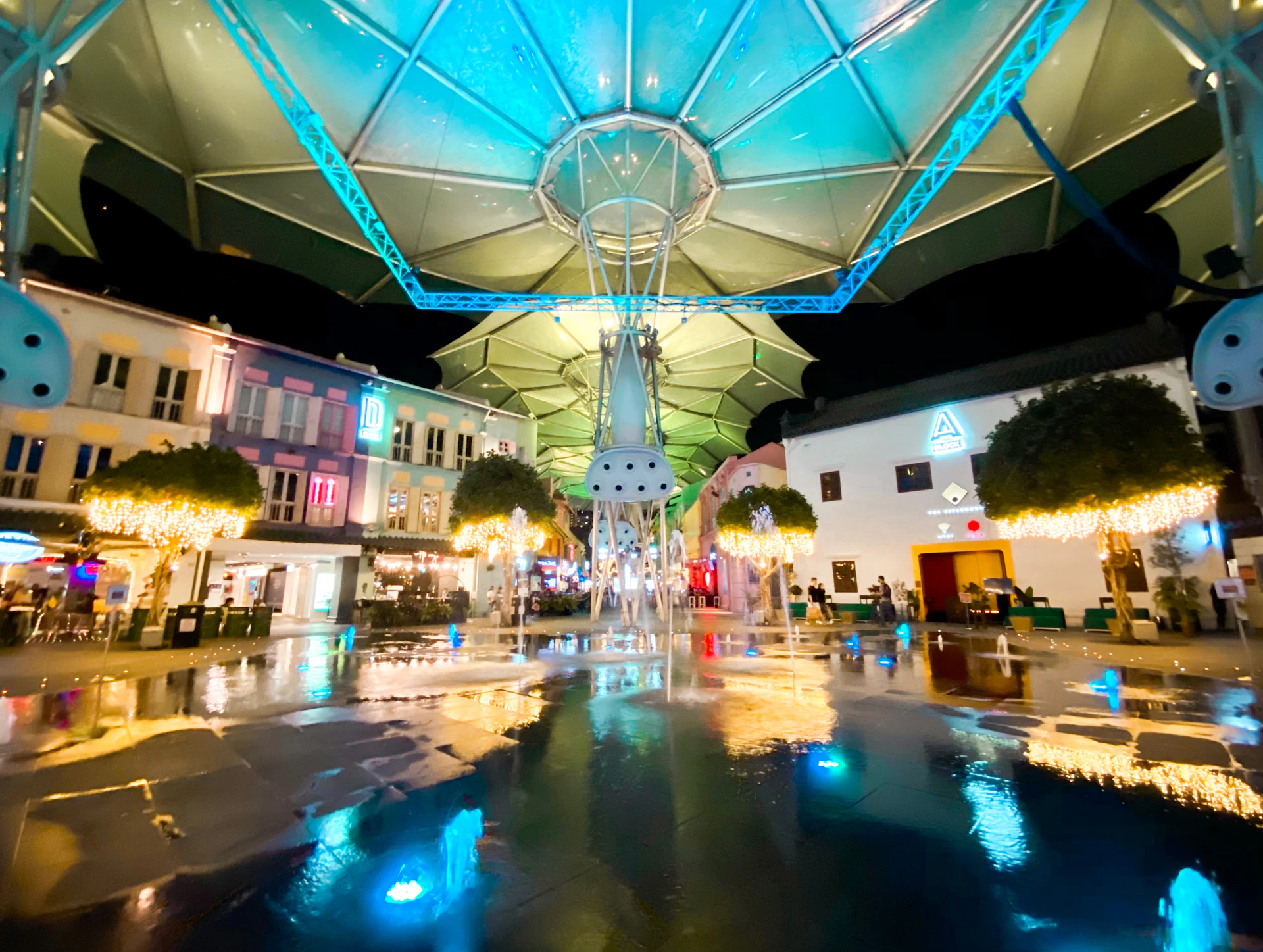 A brightly lit outdoor plaza at night with a large canopy, illuminated buildings, trees with string lights, and fountains on wet pavement.