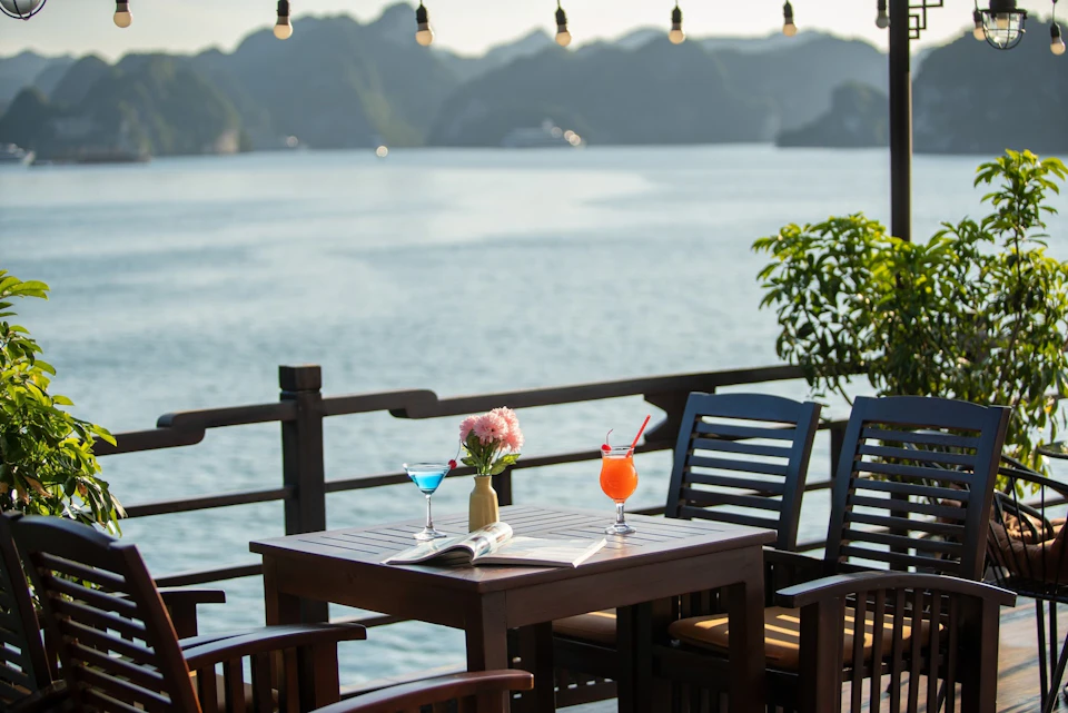 Outdoor table with two colorful drinks, a magazine, and a vase of pink flowers, overlooking a calm sea with mountainous islands.