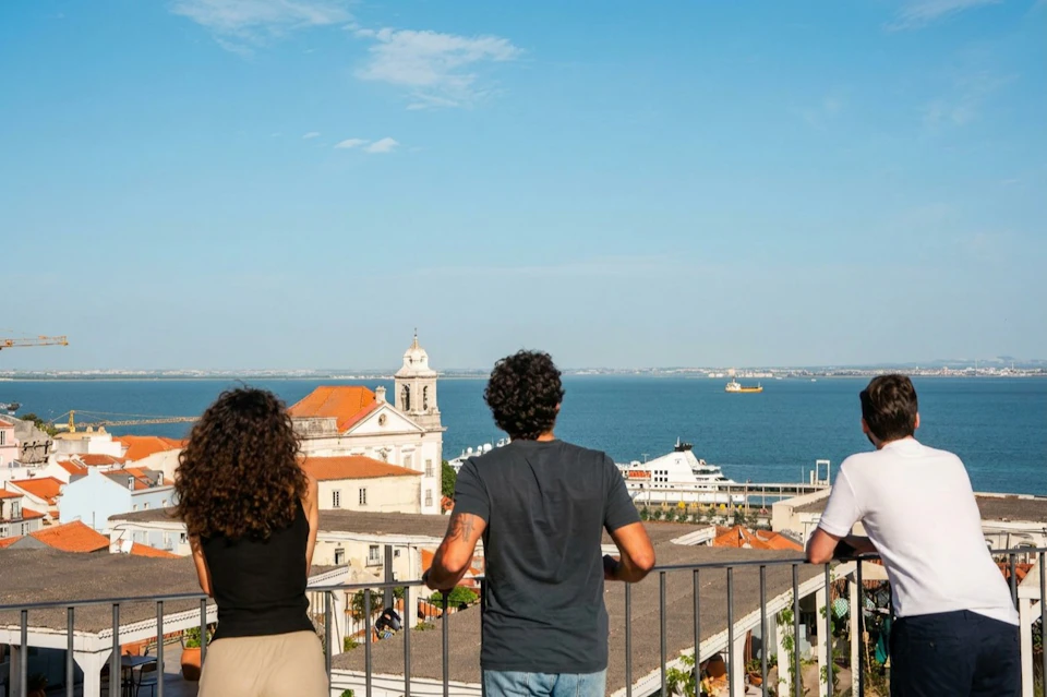Three people standing on a terrace overlooking a waterfront cityscape with a church and a ship in the distance.
