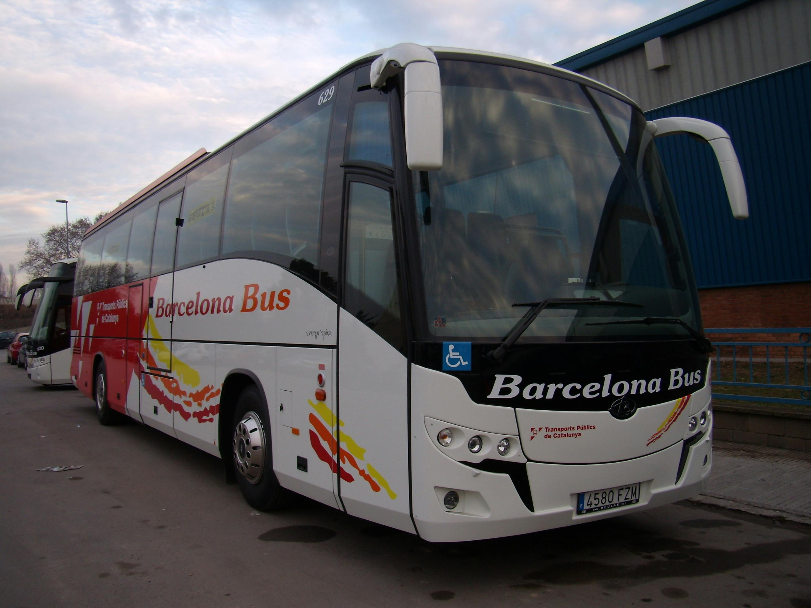 En vit och röd buss märkt "Barcelona Bus" parkerad på en asfalterad yta nära en blå industribyggnad.