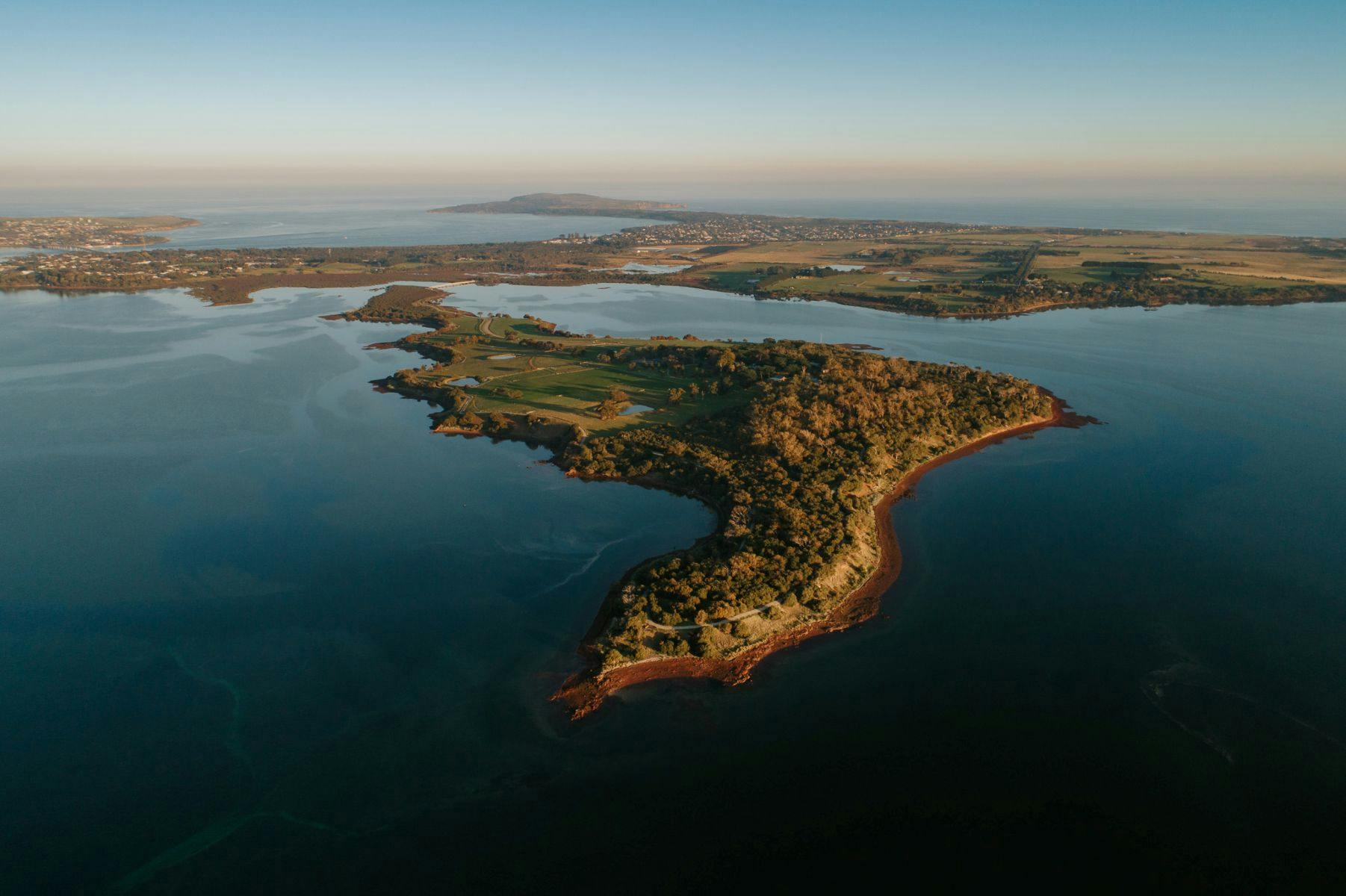Vue aérienne d'un paysage côtier comprenant une péninsule avec des champs, des arbres et des eaux calmes environnantes sous un ciel clair.