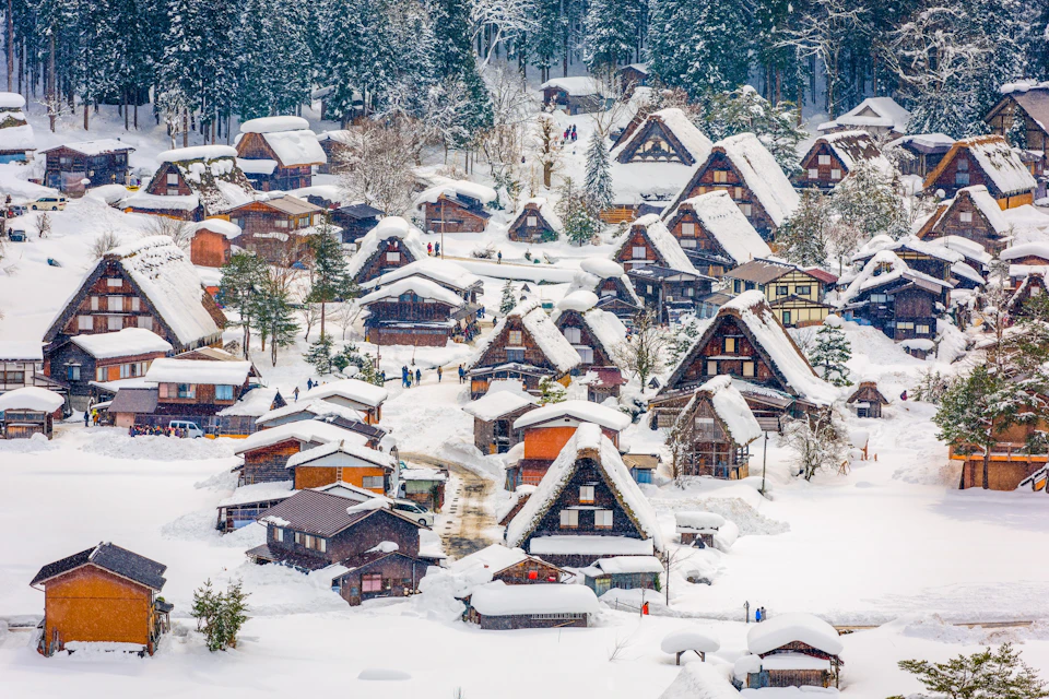 Snow-covered traditional houses in a village surrounded by trees, with people walking on paths between the buildings.