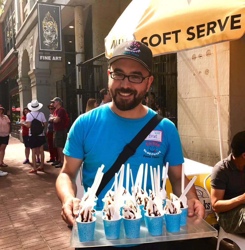 Mężczyzna uśmiecha się, trzymając tacę niebieskich filiżanek wypełnionych miękkimi lodami z sosem czekoladowym i białymi łyżkami.