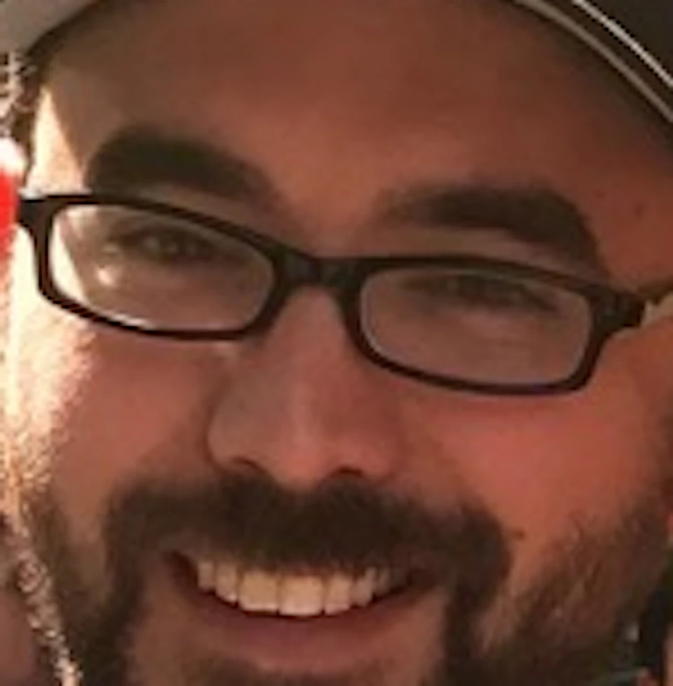 A man smiles while holding a tray of blue cups filled with soft serve ice cream topped with chocolate sauce and white spoons.