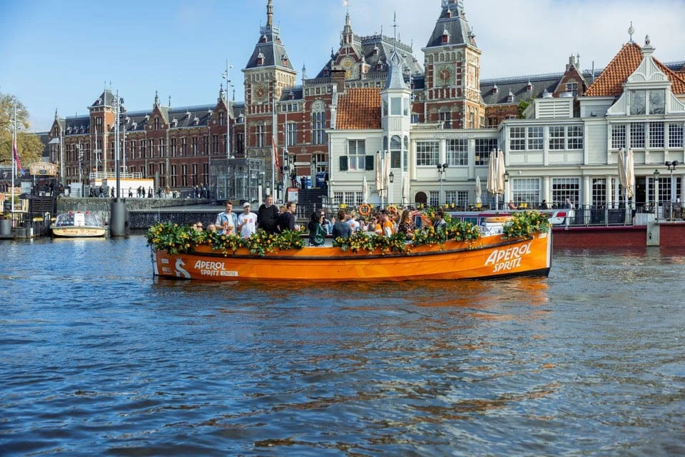 A boat decorated with flowers and the Aperol branding, filled with people, floats on a canal with historic buildings in the background.