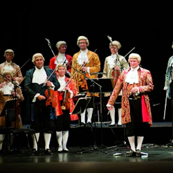 En orkester klädd i 1700-talsdräkter, stående med stränginstrument på en scen med musikställ och mikrofoner.