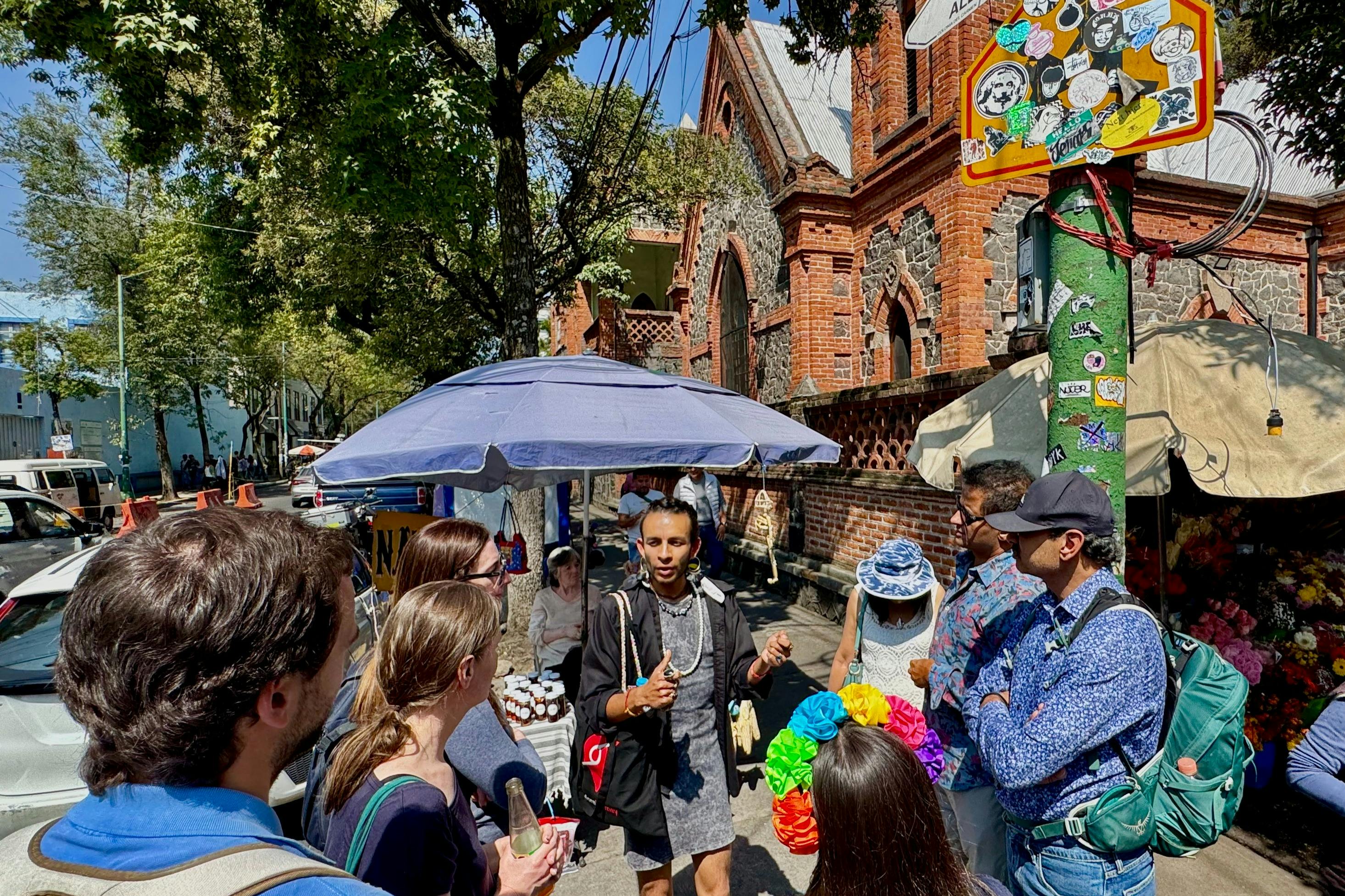 Eine Gruppe von Menschen hat sich an einem sonnigen Tag im Freien in der Nähe eines Backsteingebäudes versammelt, wobei eine Person unter einem blauen Regenschirm spricht.