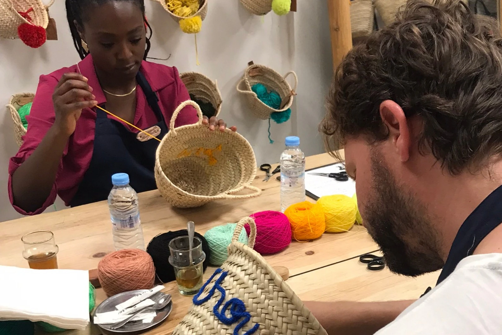 Woman painting a wicker basket at a table with yarn, water bottles, drinks, and painting supplies. Various baskets on wall.