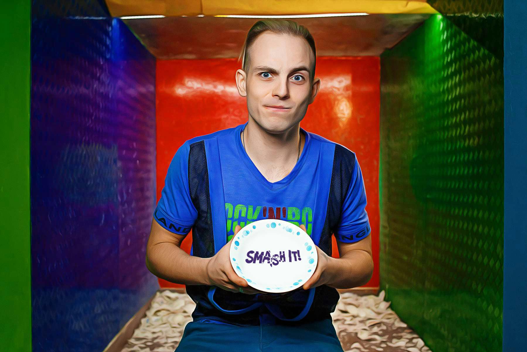 Una persona con una camicia blu tiene in mano un piatto bianco con la scritta "SMASH IT!", in piedi in una stanza colorata.