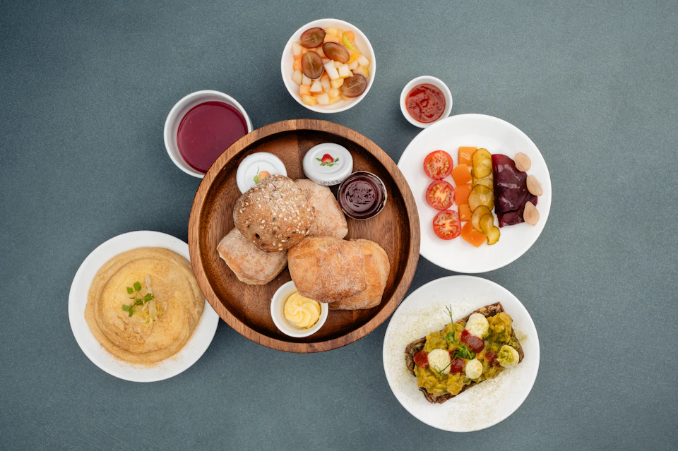 Assorted dishes on a table, including bread rolls, hummus, grilled vegetables, fruit salad, sauces, and an open-faced sandwich.