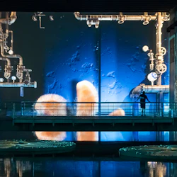 En person står på en gångväg inuti en industriell byggnad med rör och en stor skärm som visar undervattensbilder.