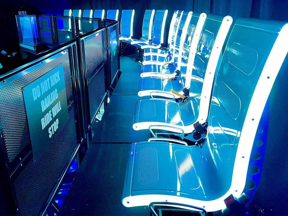 Rows of illuminated blue seats with black railings nearby, featuring a sign that reads "DO NOT KICK RAILING, RIDE WILL STOP."