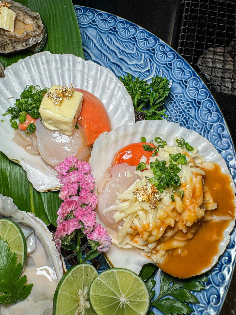 Two scallop dishes served in shells with toppings like cheese, butter, herbs, and sauce, garnished with lime and flowers.