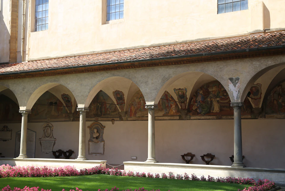 Courtyard with arched colonnade, frescoes on walls, mowed lawn, and blooming pink flowers in the foreground.