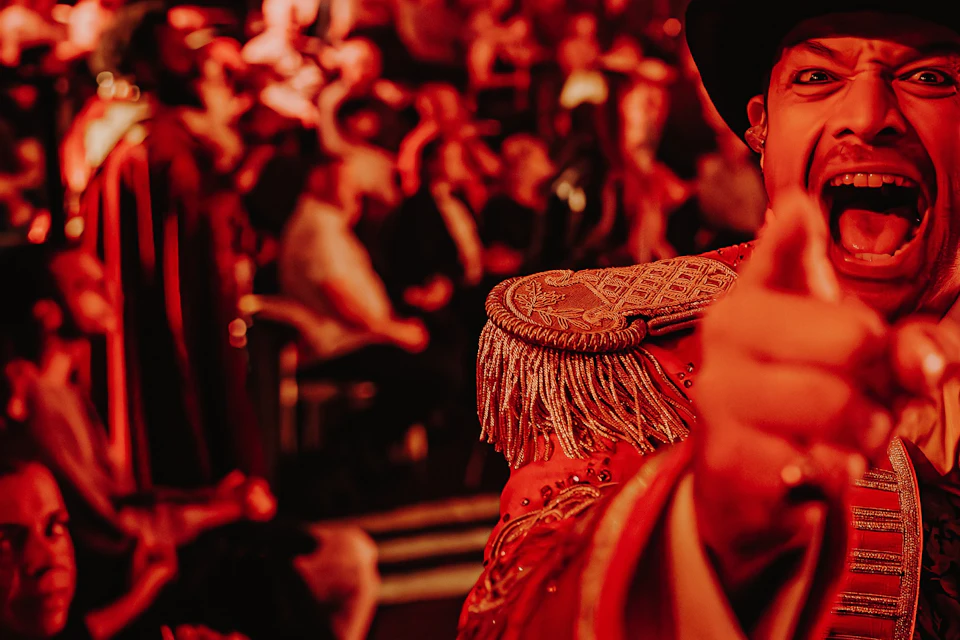 A circus ringmaster in an ornate outfit enthusiastically gestures towards the camera, with a crowd and lights in the background.